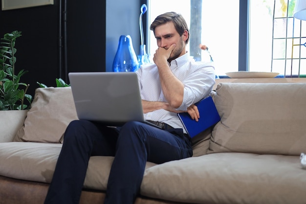 Несчастный разочарованный молодой мужчина, держащий голову за руку, сидящий с ноутбуком на диване
