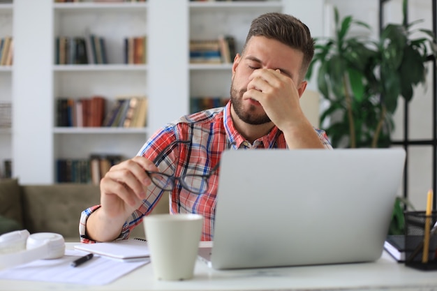 Unhappy frustrated young male holding head by hands sitting with laptop behind desk at home.