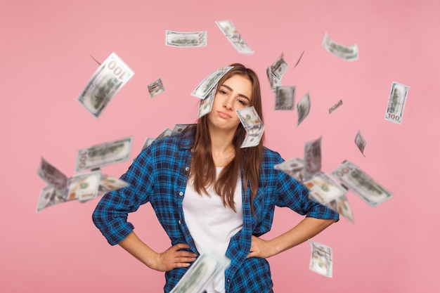 Unhappy girl in checkered shirt holding hands on hips and looking at camera with upset tired expression while money falling around worried about financial crisis debts indoor studio shot isolated