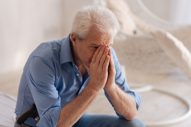 Unhappy grey haired senior man covering his face and crying while not being able to hold his feelings