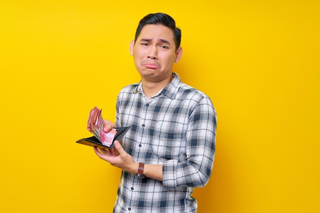 Unhappy handsome young asian man wearing a white checkered shirt holding wallet with cash money isolated over yellow background people lifestyle concept