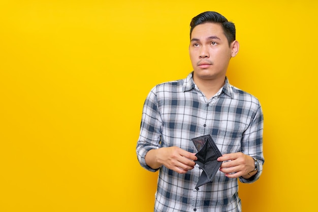 Unhappy handsome young asian man wearing a white checkered shirt showing an empty wallet and having financial problems looking side isolated over yellow background people lifestyle concept