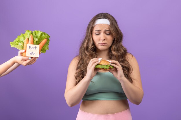 Photo unhappy hungry young european plus size female in sportswear looks at burger