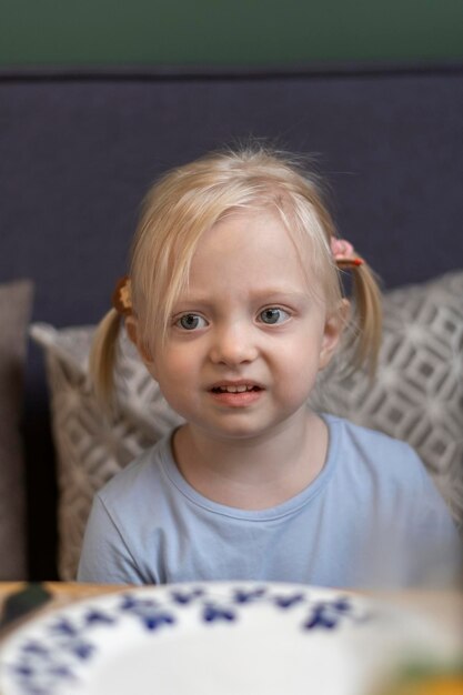 Unhappy little blonde girl with two tails sitting with empty plate at the table in cafe Hungry threeyearold girl Waiting for meal