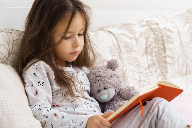 Unhappy little girl in sleepwear sit on couch and read book Portrait of child preschooler Upset kid study at home