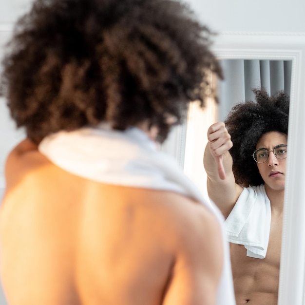 Unhappy man Body shaming Low selfesteem Dissatisfied handsome shirtless guy in eyeglasses long brown curly hair towel on bare shoulders showing thumb down gesture to reflection in mirror