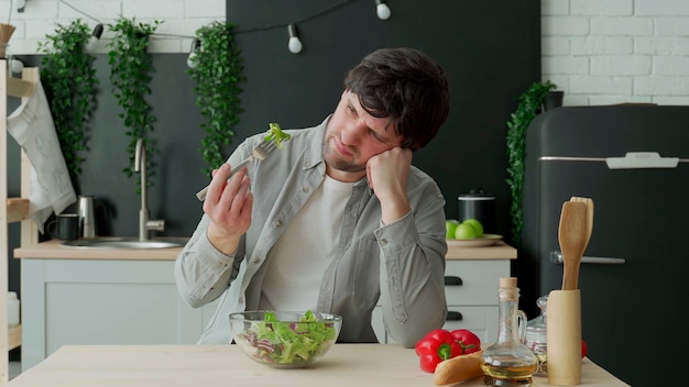 Uomo infelice che mangia insalata di verdure al tavolo in cucina