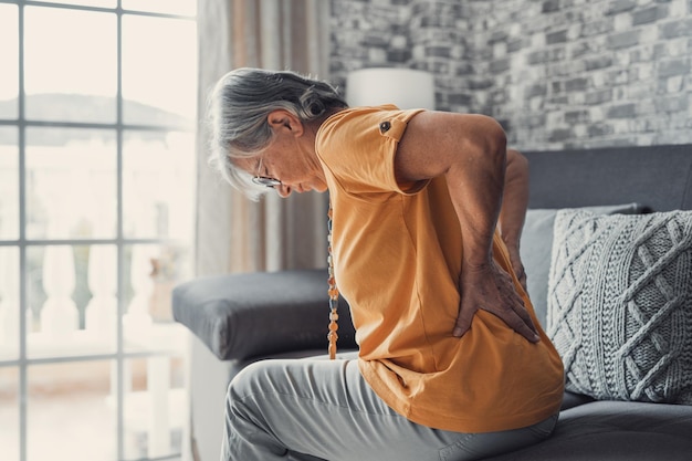 Фото Несчастная зрелая женщина, касающаяся спины, чувствующая боль, сидящая на диване в гостиной, нездоровый средний возраст.