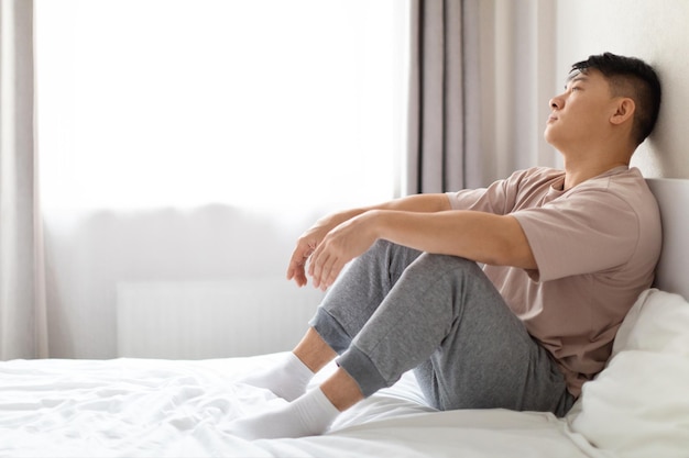 Unhappy middle aged asian man sitting on bed alone at home