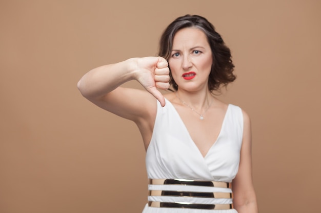 Unhappy middle aged woman showing dislike sign. Emotional expressing woman in white dress, red lips and dark curly hairstyle. Studio shot, indoor, isolated on beige or light brown background