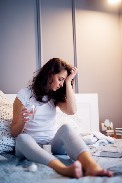 Unhappy middle aged women sitting on the bed with illness while holding a glass of water