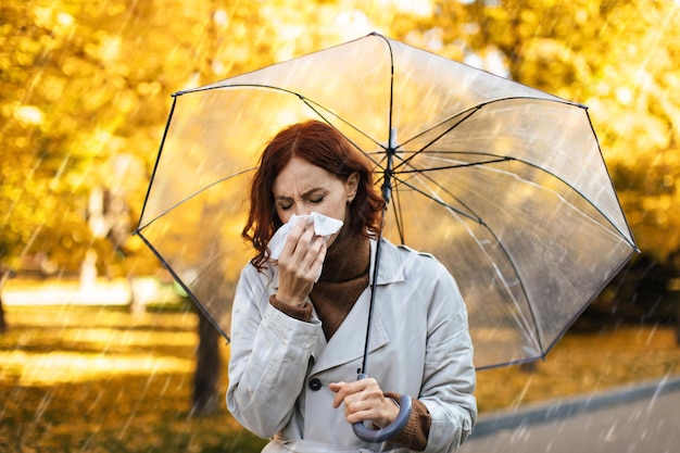 Unhappy millennial european woman in raincoat with umbrella blows her nose in napkin feels bad in park