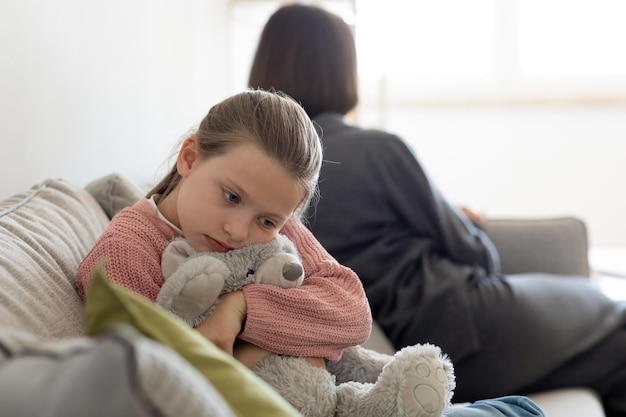 おもちゃを抱きしめる不幸な気分を害したヨーロッパの10代の女の子は、その後リビングルームのインテリアでミレニアル世代のお母さんを無視します