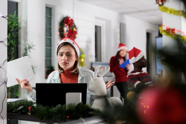 Foto impiegato infelice oberato di lavoro stressato dalle richieste di scartoffie durante le festività natalizie. donna d'affari frustrata sopraffatta dalla scadenza del progetto incombente in ufficio decorato festivo