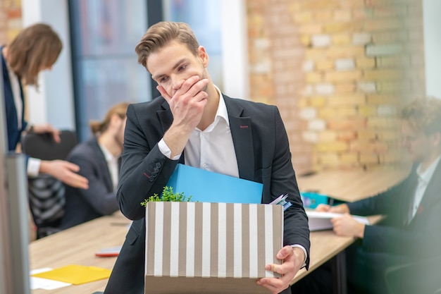 Несчастный задумчивый молодой взрослый человек в костюме, касаясь лица одной рукой, держащей коробку с вещами в офисе