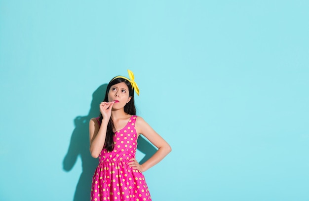 unhappy pretty girl standing in blue background eating lollipop candy and looking at empty area thinking confused problem.