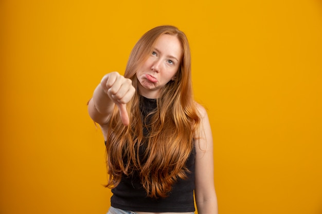 Photo unhappy redhead woman giving thumbs down gesture looking with negative expression and disapproval. yellow wall.