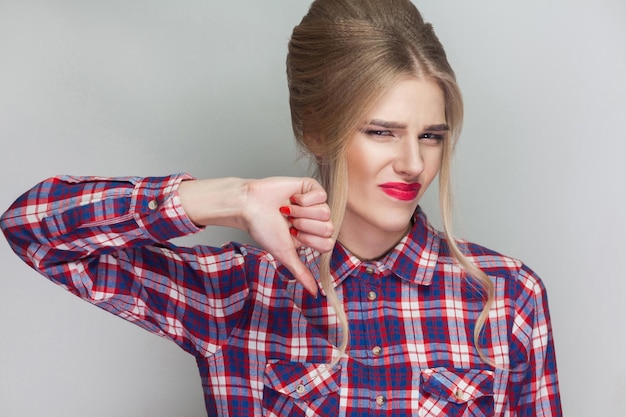 Unhappy sad girl with pink checkered shirt, collected updo hairstyle and makeup standing and looking at camera with unsatisfied face and thumbs down. indoor studio shot, isolated on gray background.