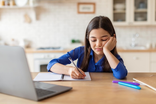 Unhappy schoolgirl taking notes at laptop tired of homework indoors