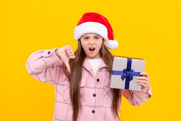 Unhappy teen girl in santa claus hat dislike present box on yellow background boxing day