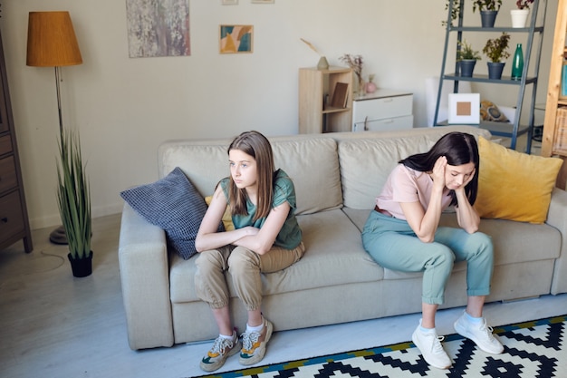 Unhappy teenage girl with crossed arms and her stressed mother with her head in hands sitting on couch during quarrel or conflict