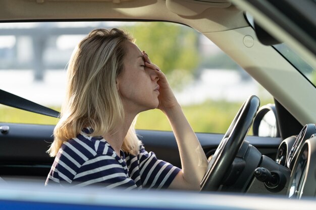 Фото Несчастная усталая водительница касается носа и лба сидит в машине и страдает от головной боли