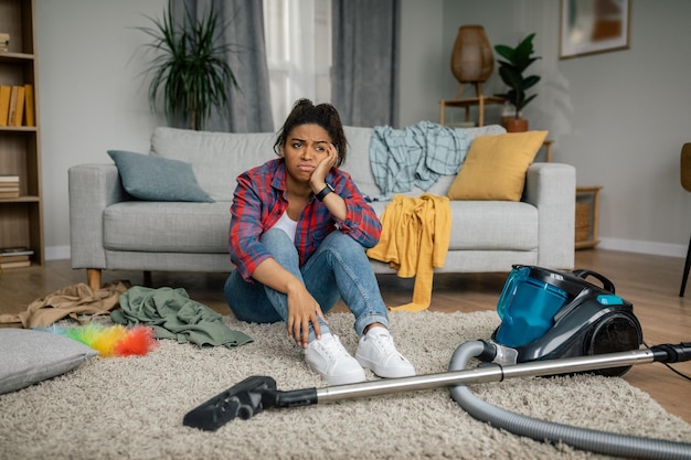 Unhappy tired young african american female sit on floor with vacuum cleaner have problems with hygiene