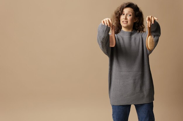 Unhappy upset curly beautiful lady in gray casual sweater with\
hairbrush comb can not make decision posing isolated on over beige\
pastel background problematic unruly damaged hair concept copy\
space