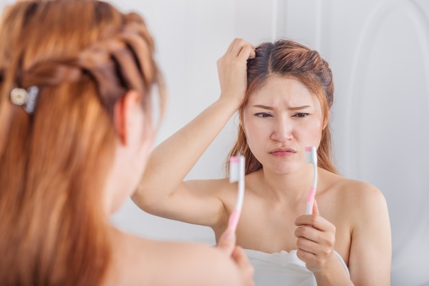 unhappy woman in bath towel brushing teeth with mirror in bathroom