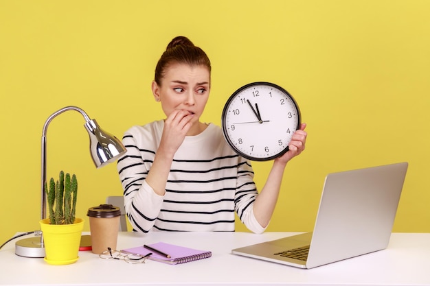 Unhappy woman office worker biting finger nails holding wall clock talking video call on laptop