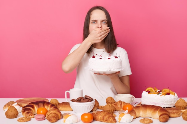 Unhappy woman wearing white Tshirt sitting at festive table with various desserts holding cake in hand covering mouth with palm being shocked or felling nausea isolated over pink background