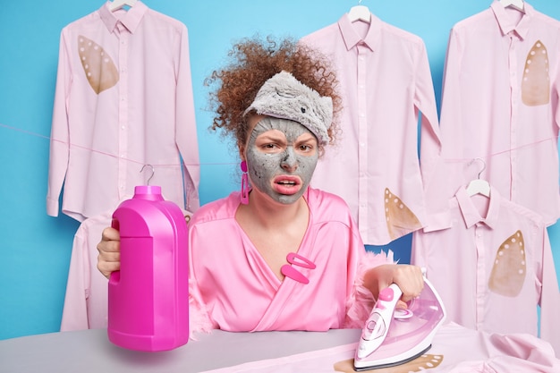 Photo unhappy woman with curly hair dressed in domestic clothes holds detergent bottle uses electric iron for stroking clothes undergoes beauty procedures. dissatisfied housewife tired of housework