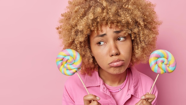 写真 巻き毛の不幸な女性は2つのカラフルなロリポップを持っています悲しい表情はダイエット財布を保ちます唇はあなたの昇進のためにピンクの背景の空のスペースの上に隔離されたファッショナブルなジャケットを着ています