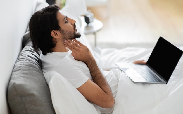 Unhappy young arab guy in white tshirt sits on bed checks tonsils has video call on laptop with