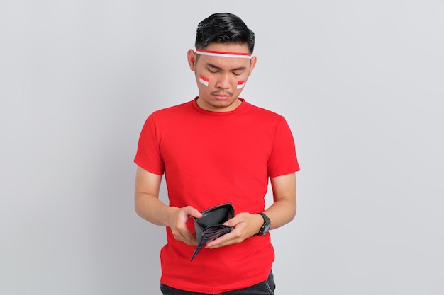 Unhappy young Asian man celebrating Indonesian independence day while looking into empty wallet isolated on white background