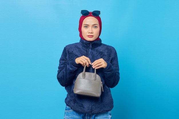 Unhappy young Asian woman holding bag and putting glasses on head on blue background