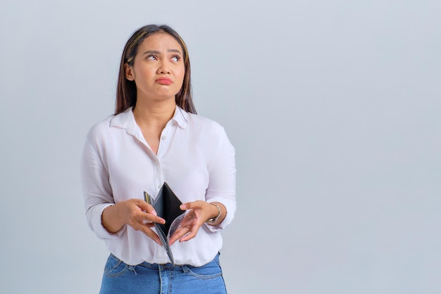 空の財布を持ち、白い背景に分離されたコピー スペースを見て不幸な若いアジア女性