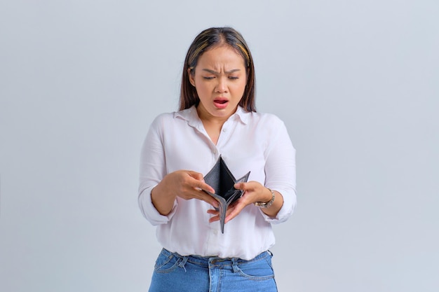 Unhappy young Asian woman looking into empty wallet isolated over white background