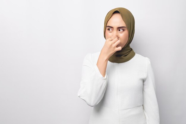 Unhappy young beautiful Asian Muslim woman 20s wearing hijab feeling disgusted holding her nose to avoid smelling foul looking aside isolated on white background People religious lifestyle concept