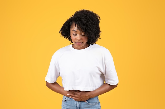 Unhappy young black curly lady in white tshirt presses hands to belly suffers from stomach pain