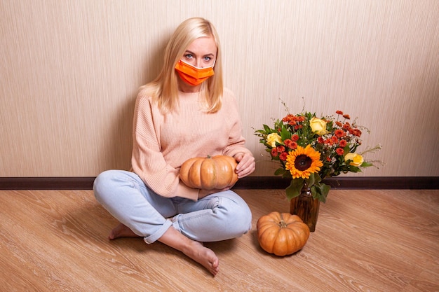 Unhappy young blonde woman wearing medical mask in warm sweater is sitting on the floor and holding a pumpkin in her hands, autumn bouquet with a sunflower nearby.