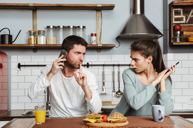 Unhappy young couple sitting at the kitchen during lunch at home, having a problem, holding mobile phone