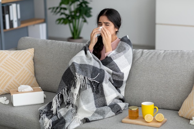 La giovane signora indiana infelice starnutisce nel tovagliolo soffia il naso soffre di febbre influenza sul divano nell'interno della stanza con il tè