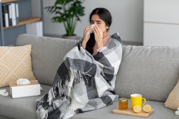 Несчастная молодая индийская леди чихает в салфетку, сморкается, страдает лихорадочным гриппом на диване в интерьере комнаты с чаем