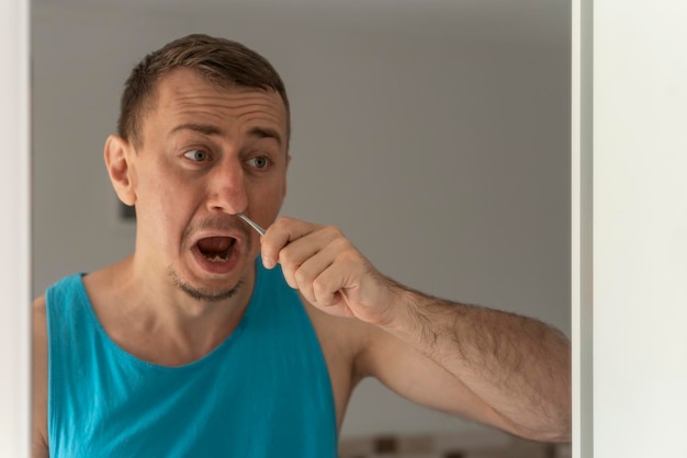 Photo unhappy young man in blue tshirt removing unwanted hair in nose standing in front of mirror morning routine