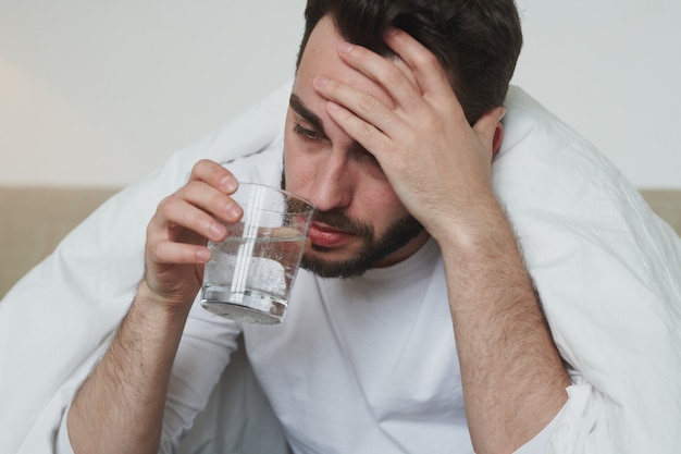 Foto giovane infelice con covid19 o influenza che si tocca la fronte e beve acqua dal vetro mentre è seduto sul letto durante l'autoisolamento a casa