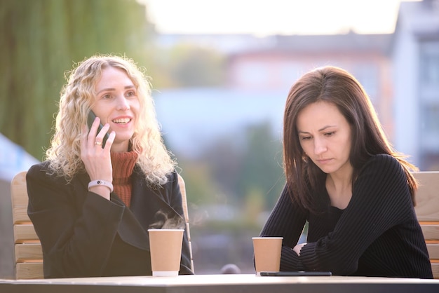Unhappy young woman waiting angrily while her friend is talking happily on sellphone with someone else and ignoring her Friendship problems concept