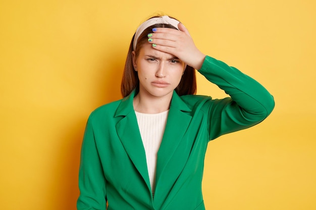 Photo unhealthy displeased woman wearing green jacket posing isolated over yellow background frowning face touching her forehead suffering high temperature