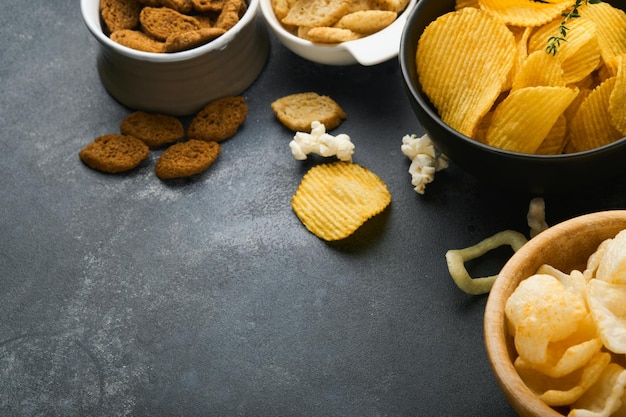 Cibo malsano snack tutti i classici snack di patate con popcorn alle arachidi e anelli di cipolla e salatini salati in piatti di ciotola su fondo di cemento nero cibo malsano per i denti della pelle del cuore della figura