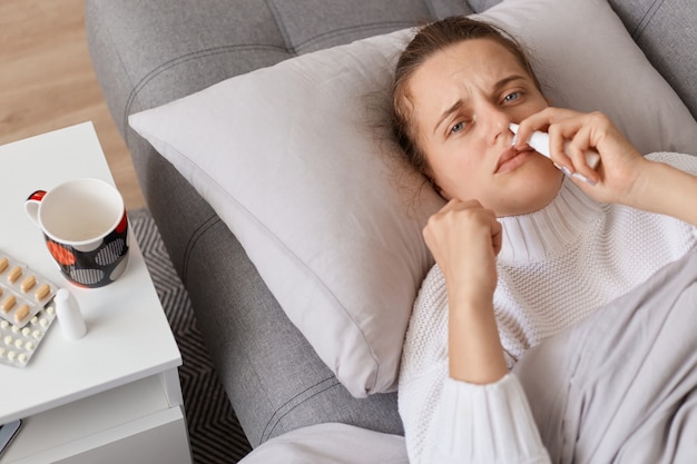 Donna malata malsana sdraiata sul divano e usando spray nasale, guardando la telecamera, trattando grippe, stare a casa, avere sintomi influenzali, ha bisogno di cure.